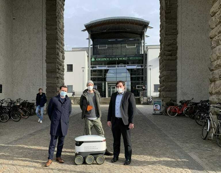 Drei Männer stehen mit kleinem weißen Roboter vor Klinikeingang